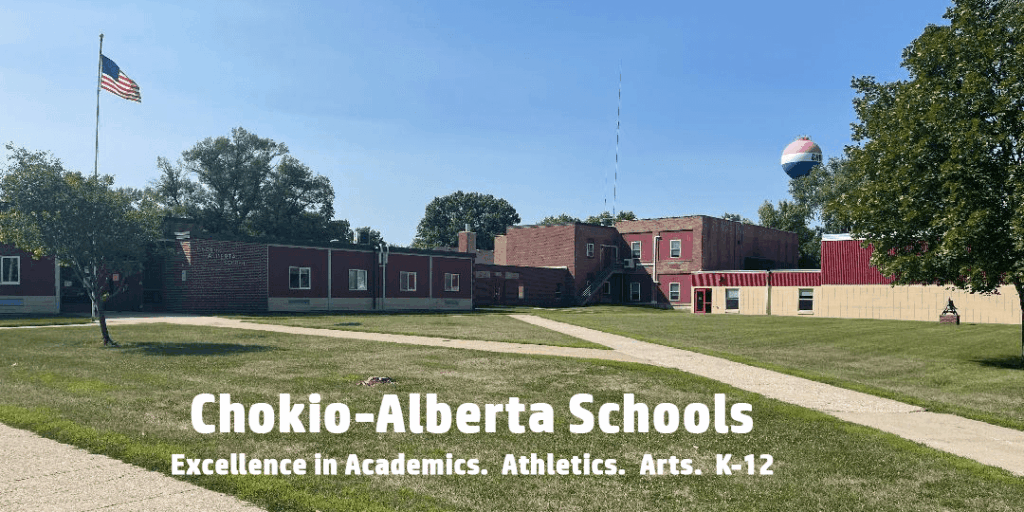 Wide outdoor view of the Chokio-Alberta Schools campus on a sunny day, showing brick school buildings, a grassy lawn with sidewalks, an American flag on a flagpole, and a water tower in the background. Text over the image reads, “Chokio-Alberta Schools – Excellence in Academics. Athletics. Arts. K-12.”