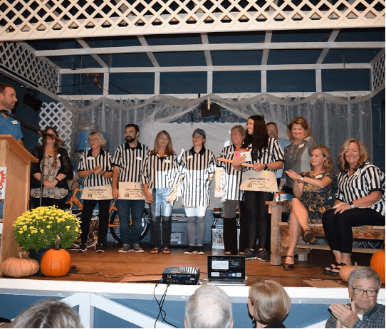 Group of adults standing on a decorated stage at an indoor event, many wearing black-and-white referee-style shirts and holding gift bags. A speaker stands at a podium on the left, while pumpkins and fall decorations sit at the front of the stage. An audience is visible in the foreground.