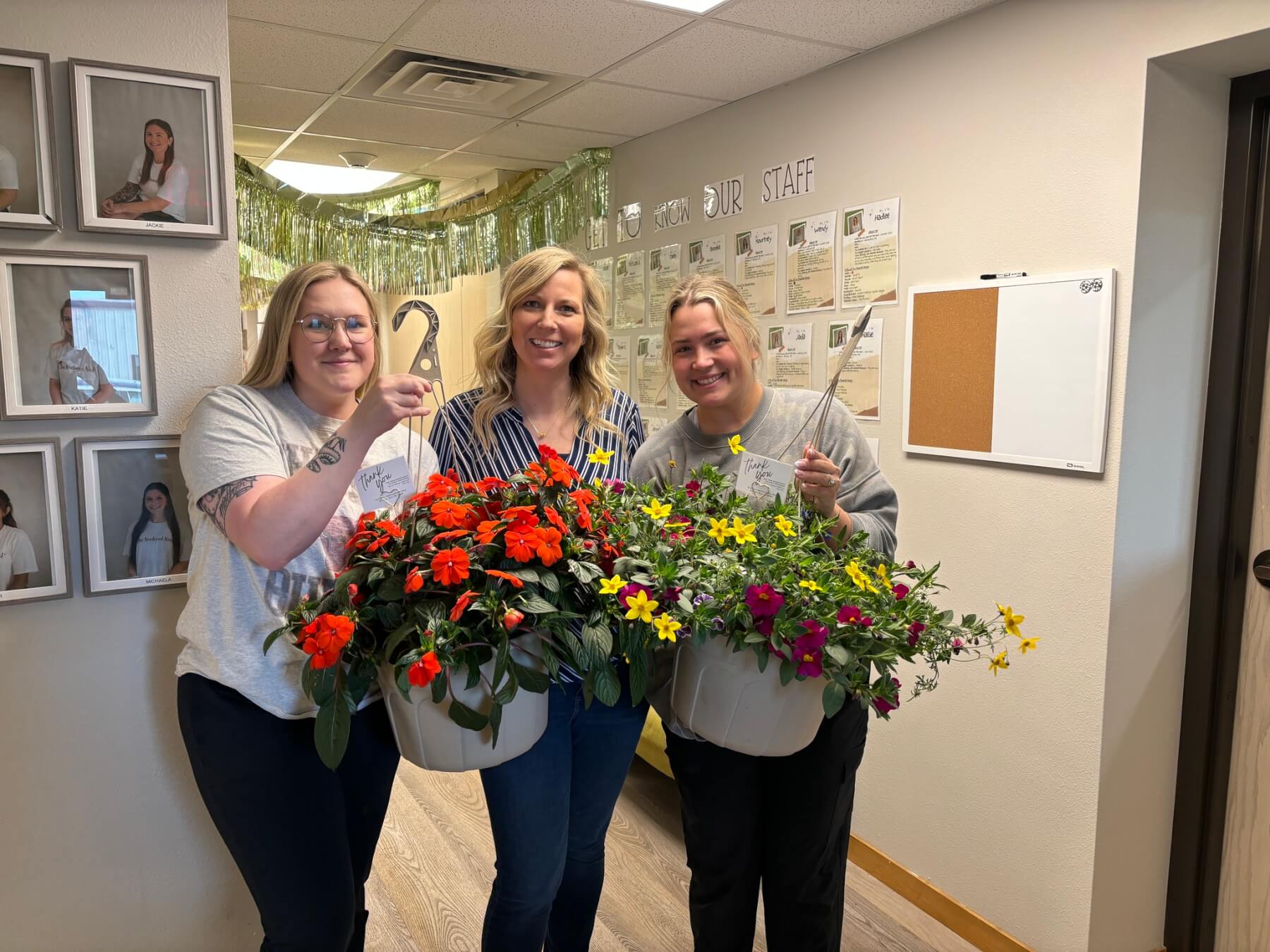 Three women standing in an office hallway holding large hanging baskets filled with colorful flowers, smiling at the camera. Framed staff photos and a bulletin board are visible on the walls behind them.