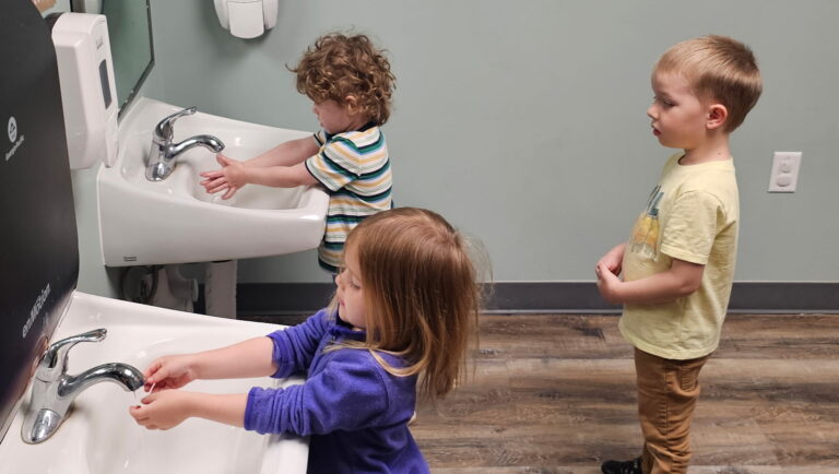 A boy and girl wash their hands in two small sinnks and one child waits to wash his hands.